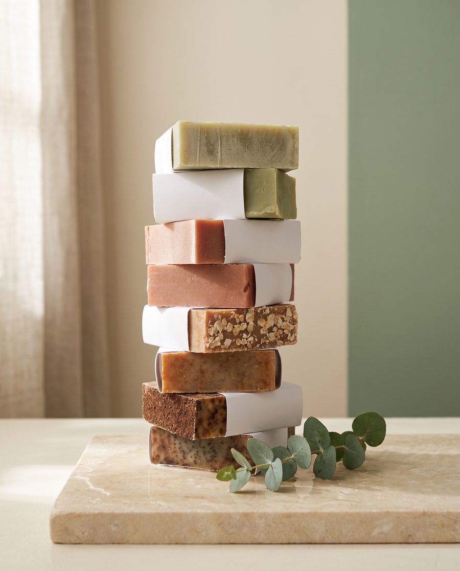 A tall stack of handmade Nested Arrow soap bars in white paper sleeves on warm ivory stone with eucalyptus